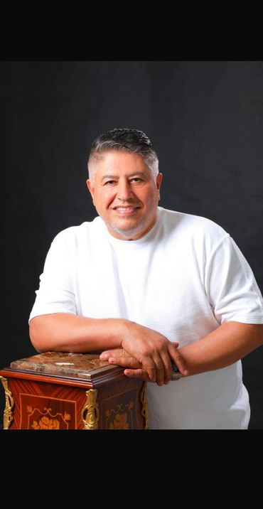 Smiling man in white shirt leaning on an ornate wooden box against a black background.