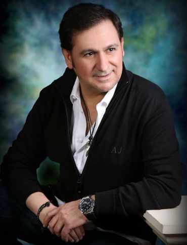 Man in a black jacket with a white shirt, smiling and sitting relaxed.