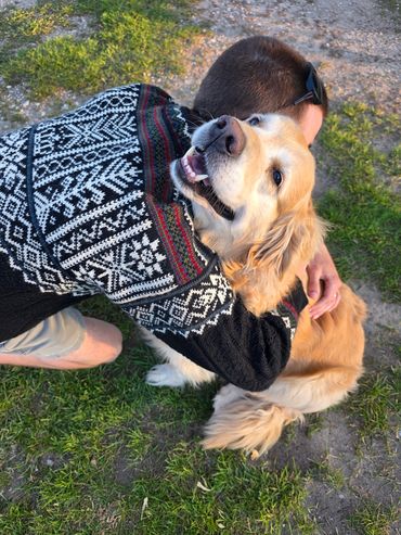 A man in a patterned sweater hugs a happy golden retriever outdoors.