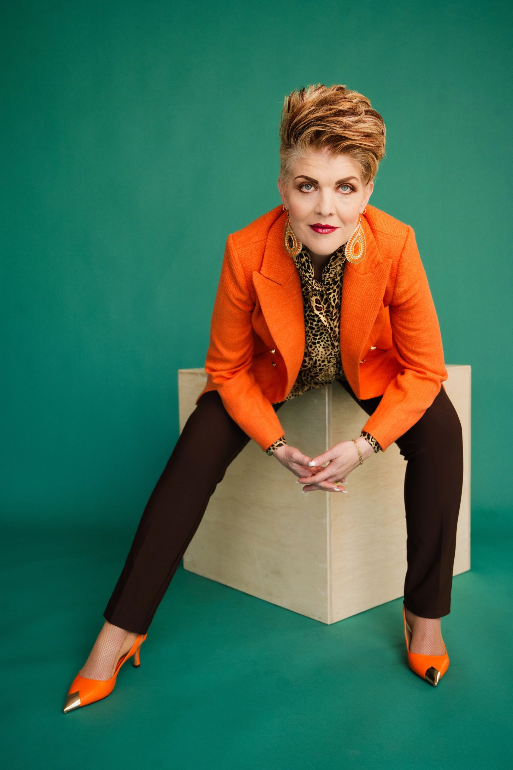 Confident woman in orange blazer and leopard print shirt poses against teal background.