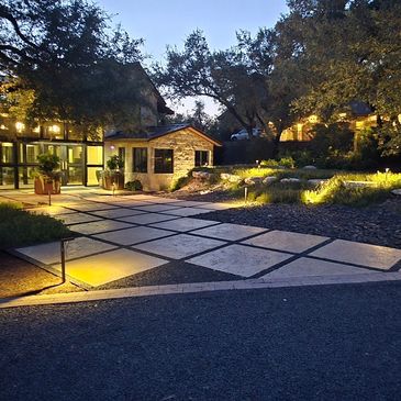 Evening view of a well-lit driveway with modern landscaping and stone buildings.