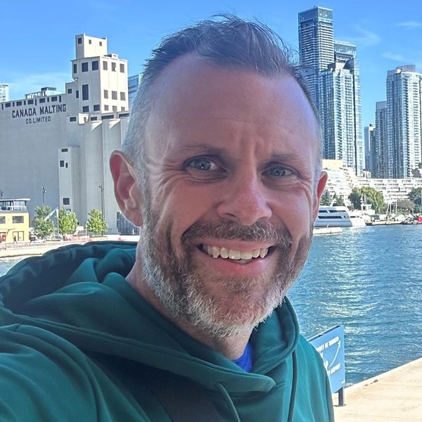 Smiling man in green hoodie by waterfront with city skyline.