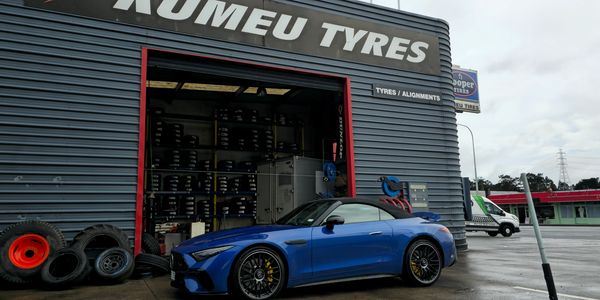 Blue sports car parked outside a tire shop with stacked tires.
