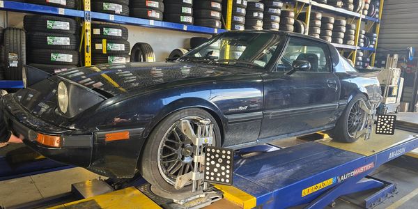 A black Mazda RX-7 sports car on a wheel alignment machine in a tire shop.