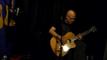 A man intensely playing an acoustic guitar on stage in a dimly lit room.