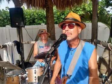 Two men playing music outdoors under a thatched roof setup.