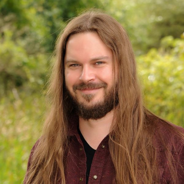 Man with long hair and beard smiling outdoors.