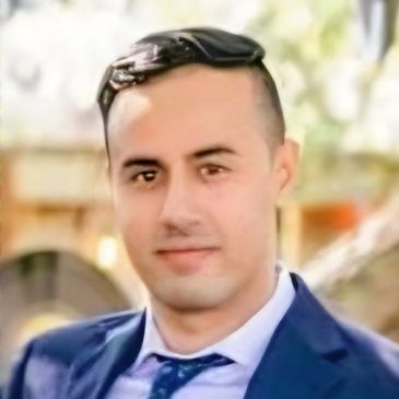 A young man in a blue suit and tie poses outdoors with a soft-focus background.