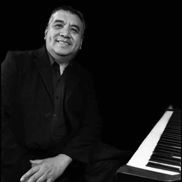 Man in black suit smiling beside a piano keyboard in a black and white photo.