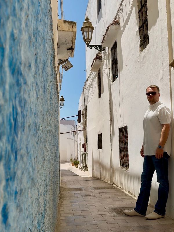 Man in sunglasses stands in a narrow alley with white and blue walls.