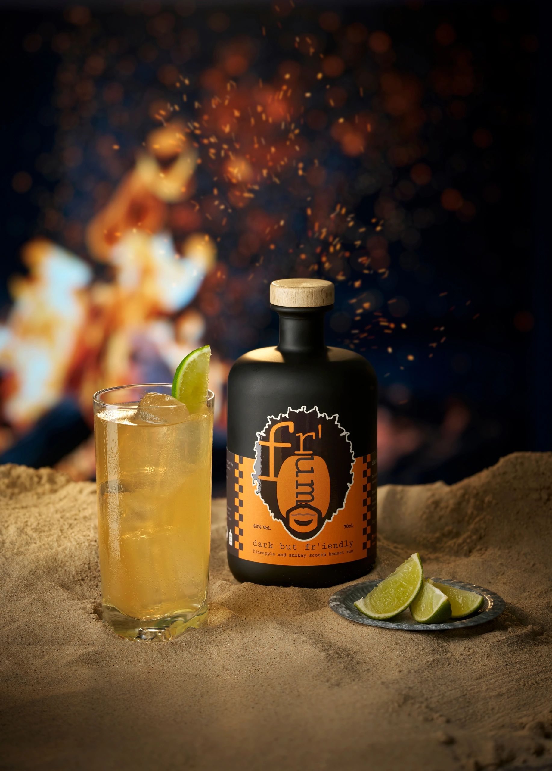 Bottle of Fr'um on the beach sat in the sand with a fire in the background
