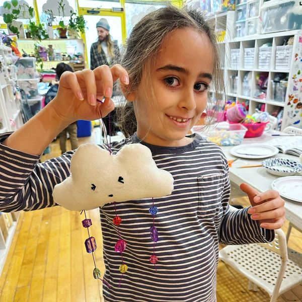 Smiling girl holding a handmade cloud craft with colorful beads in a busy art room.