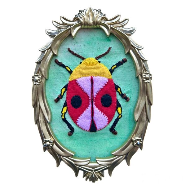 Colorful embroidered bug in an ornate gold frame on green fabric.