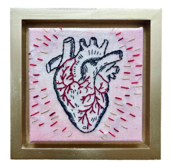 Embroidered anatomical heart with vibrant red veins on pink fabric, framed in gold.