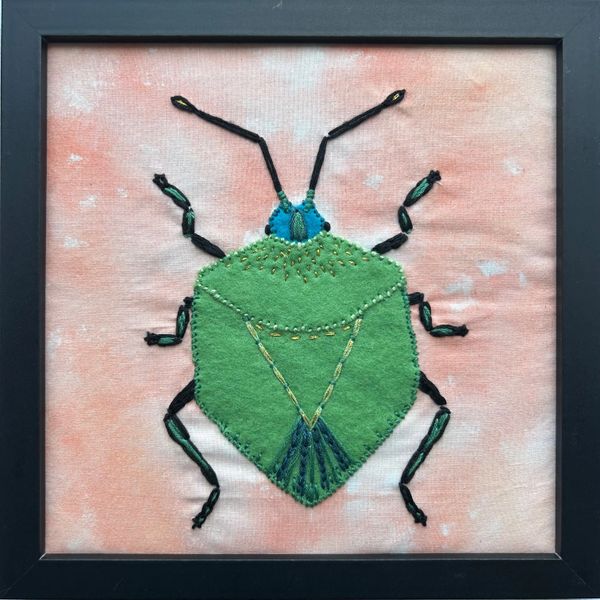 Framed felt and embroidered green bug on a peach fabric background.