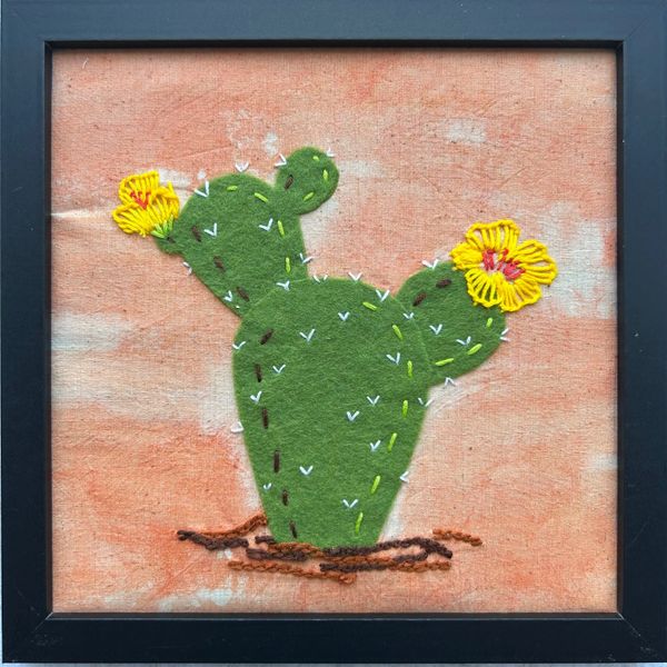 Framed embroidery of a green cactus with yellow flowers on a textured peach background.