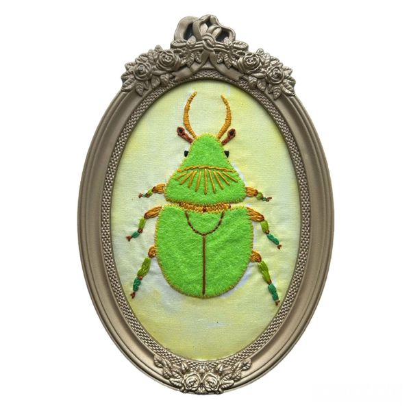 Embroidery of a green beetle in an ornate oval frame.