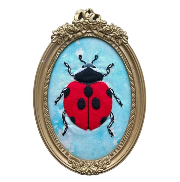 Framed embroidered ladybug on blue fabric with ornate gold frame.