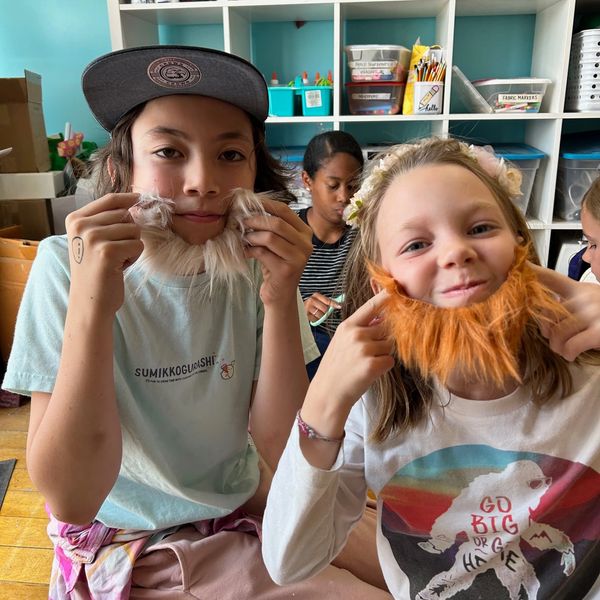 Two kids wearing fake beards and smiling in a colorful classroom.
