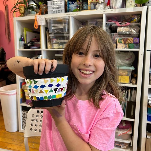 A smiling girl proudly shows her colorful mosaic pot in a creative art room.