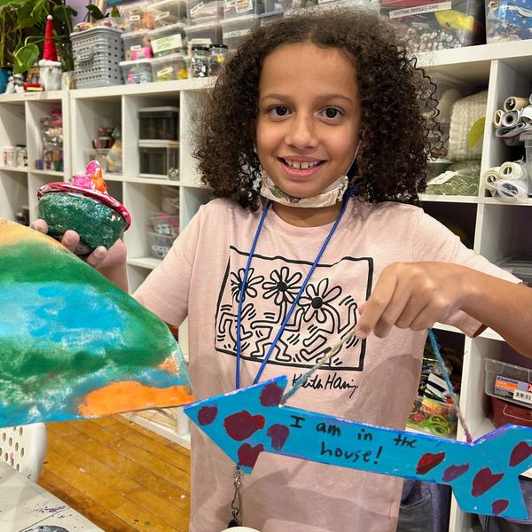 A smiling child shows colorful handmade crafts in an art room.