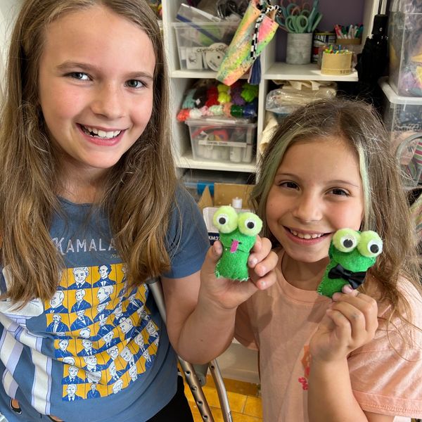 Two girls smiling and holding green felt frog crafts in a colorful craft room.