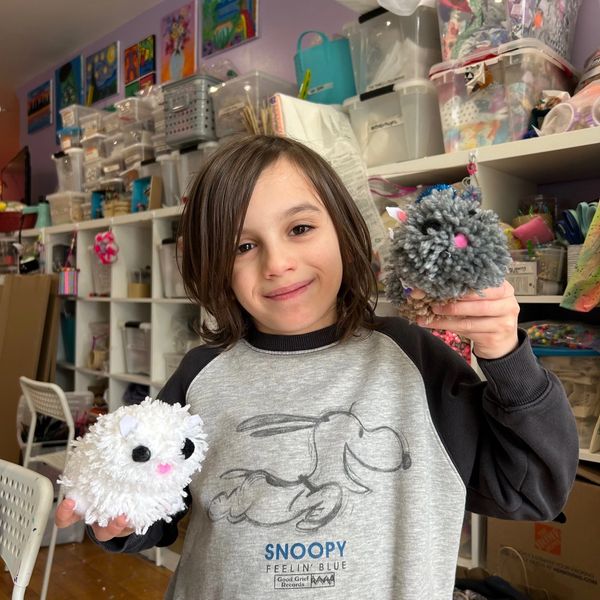 Child holding two fluffy pom-pom crafts, smiling in a colorful room.