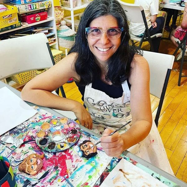 Woman smiling while painting colorful character faces at a busy art table.