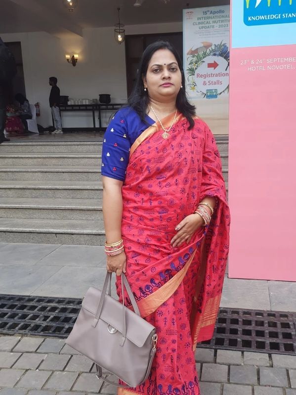 Woman in a red saree with blue blouse holding a grey handbag.
