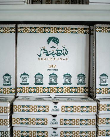 Stacked Shahbandar branded boxes with QR codes and decorative patterns.