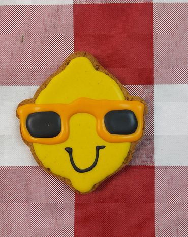 Smiling lemon-shaped cookie with orange sunglasses on a red and white checkered tablecloth.