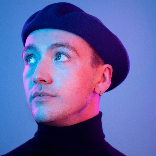 Simon wears a blue beret and turtleneck. They are facing away and are being lit under red and blue l