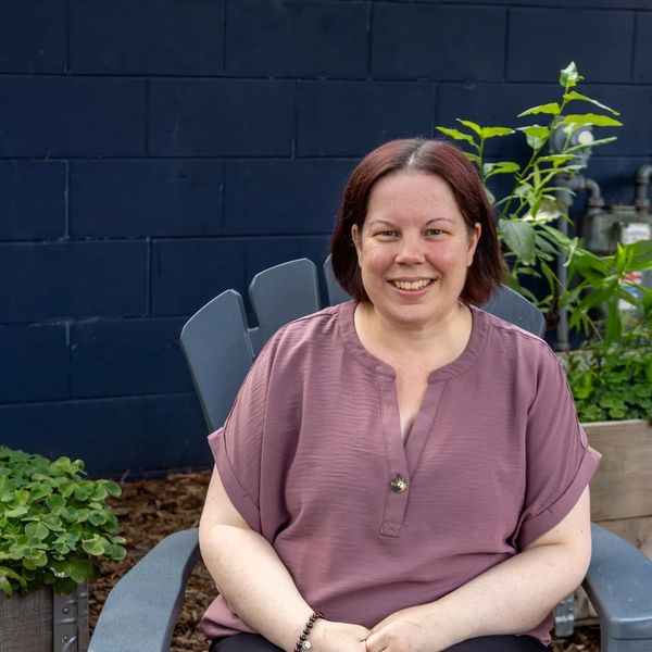 Smiling woman sitting outdoors on a gray chair with green plants in the background.