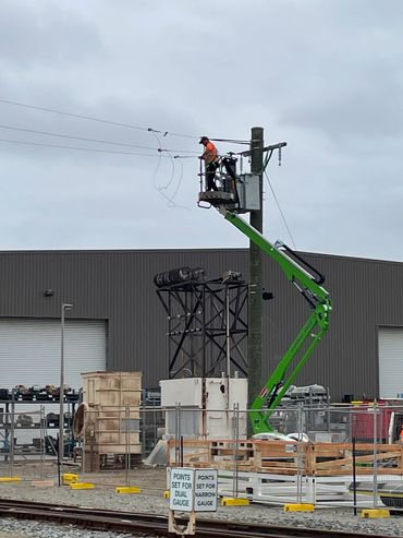 Worker in orange using a green lift to repair power lines on a pole at a construction site.
