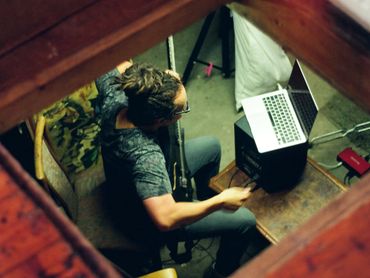 Man with dreadlocks playing guitar and using a laptop in a cozy room.