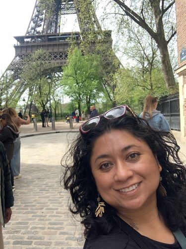 Gabriela smiling in front of the Eiffel Tower. She has short curly brown hair. 