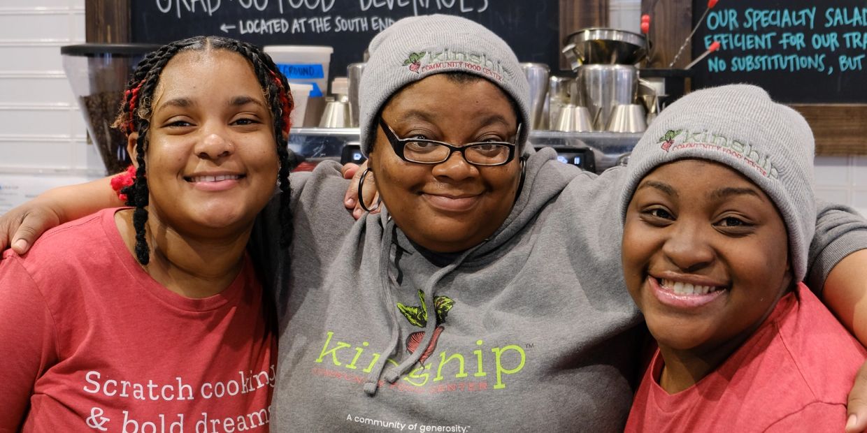 Three women smiling together indoors, wearing branded clothing.