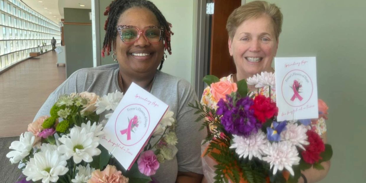 Two smiling women holding colorful flower bouquets with pink ribbon cards.