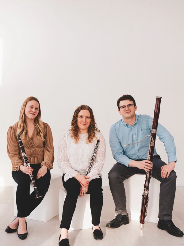 Three musicians seated holding woodwind instruments against a plain background.