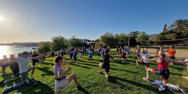 A group exercise class outdoors on a sunny day near water.