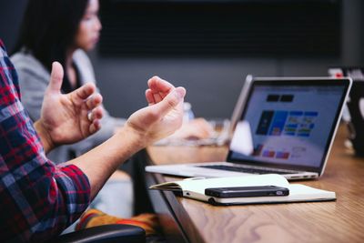 Person gesturing during a meeting with laptop and notebook on table.