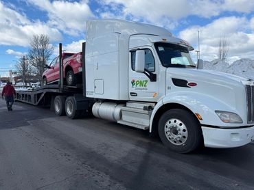 White semi-truck from PNZ Transports carrying a red pickup on a trailer under a cloudy sky.