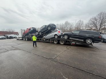 A truck transports multiple SUVs on a car carrier under a cloudy sky.