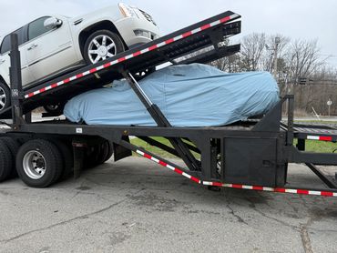 Two cars are loaded on a car carrier truck, one covered with a blue tarp.