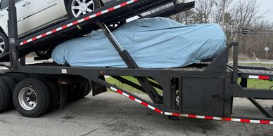Two cars are loaded on a car carrier truck, one covered with a blue tarp.