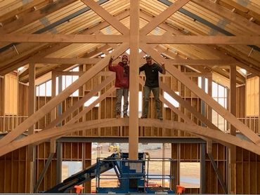 Two men standing on a wooden beam inside a timber-framed building under construction.