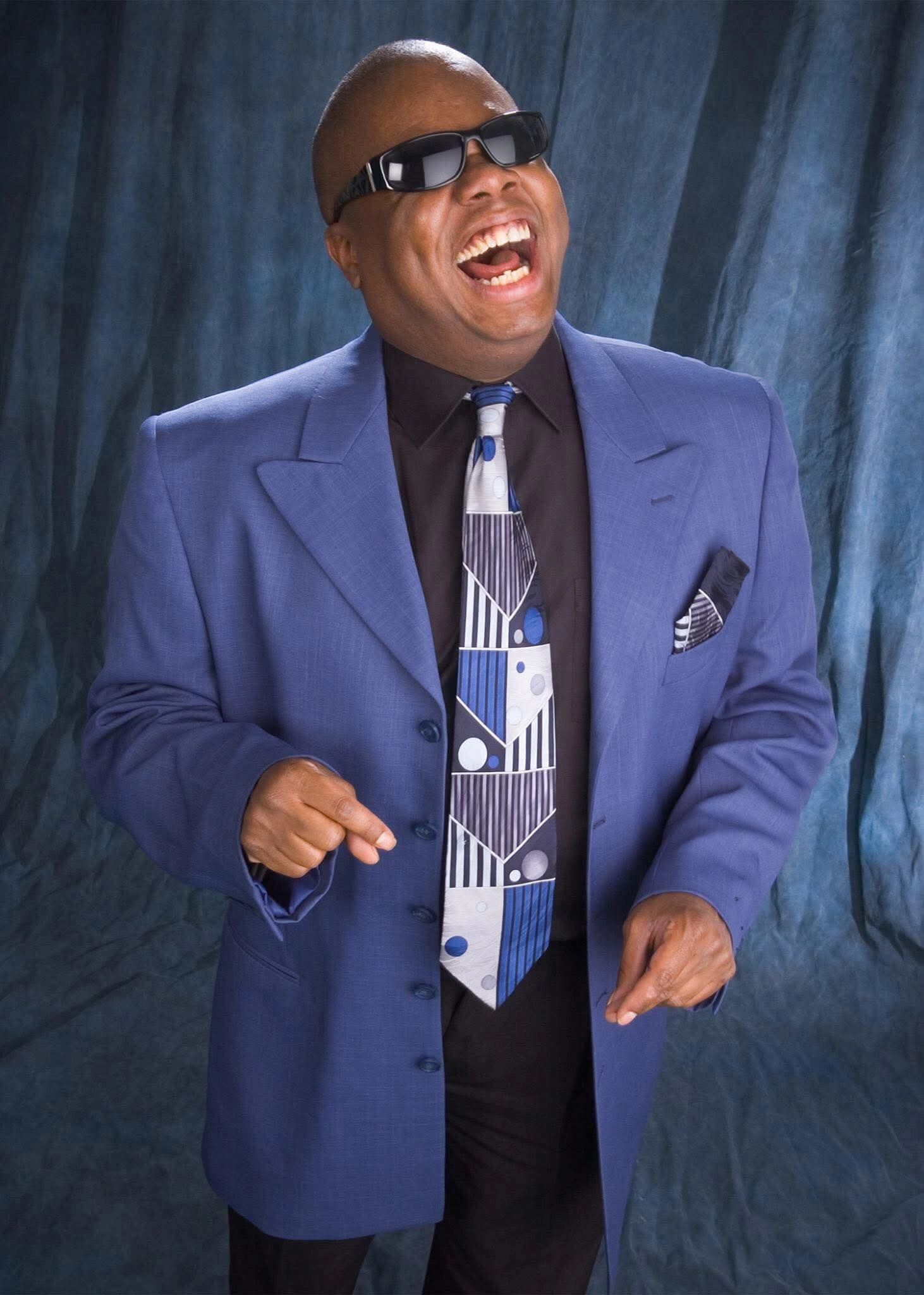 Man in blue suit and sunglasses laughing heartily.