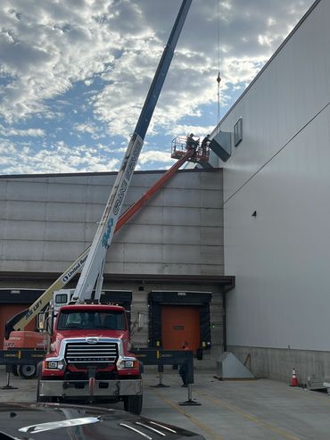 Workers use cranes to install equipment on a warehouse wall under a cloudy sky.