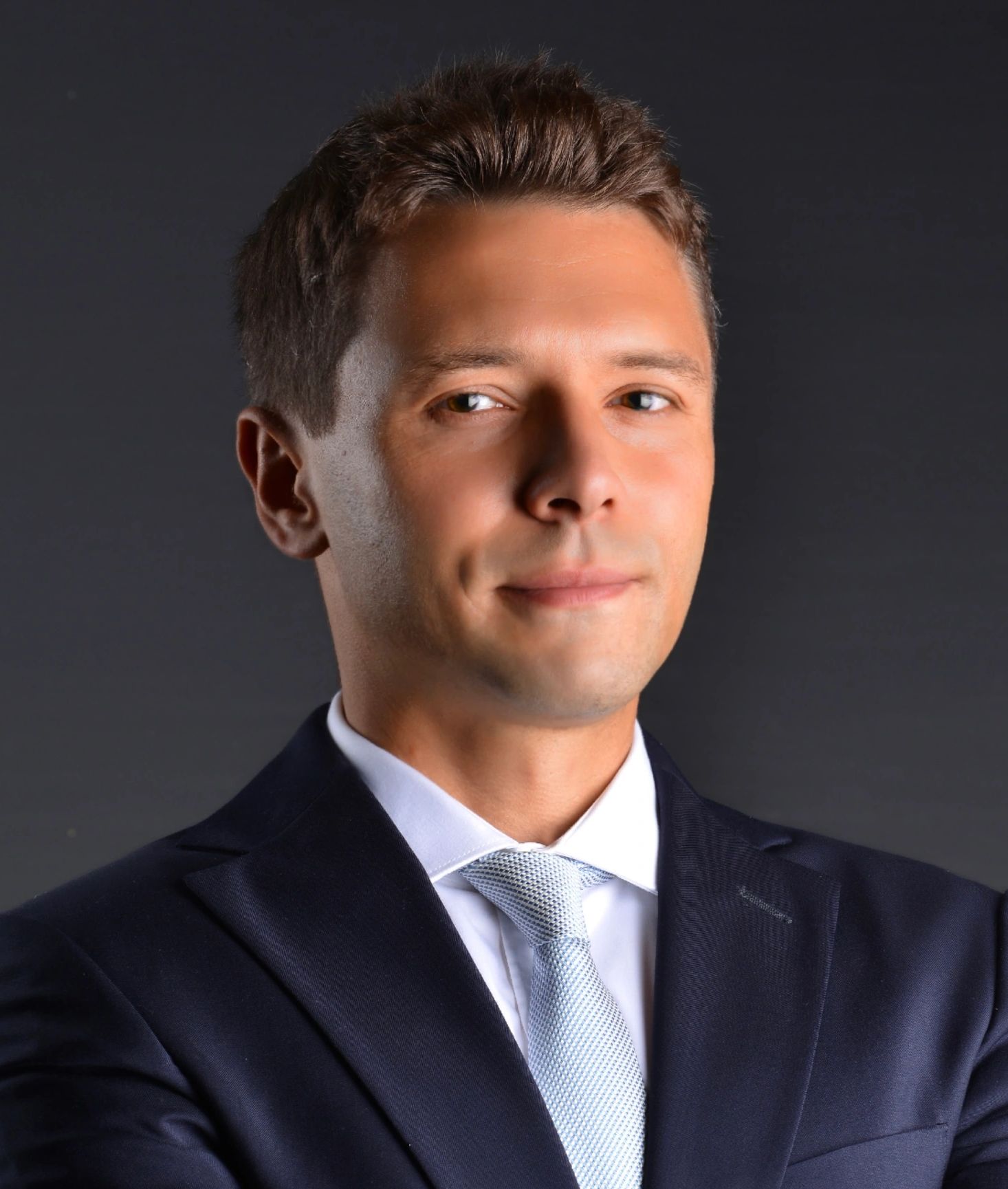 Confident man in a dark suit and light blue tie posing with crossed arms.