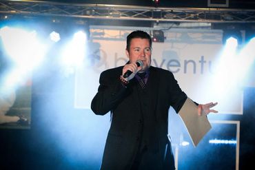 Man in suit speaking into microphone on stage with bright lights.